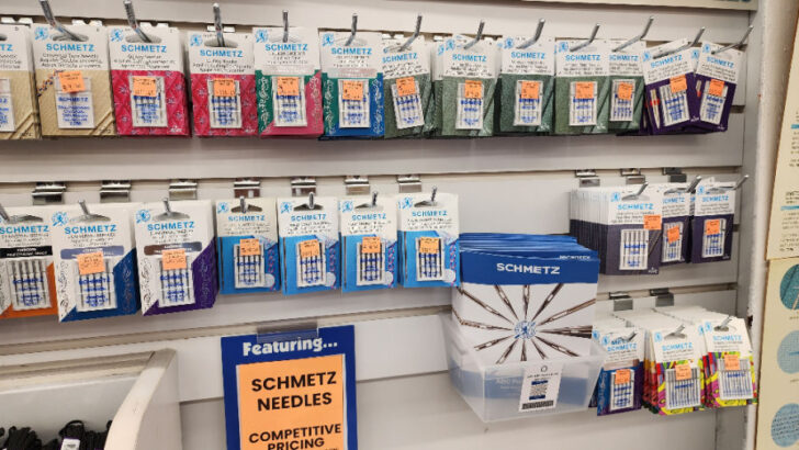 The display is neatly arranged with Schmetz needle packs hanging on hooks, sorted by category. Each pack has labels indicating the needle type, size, and intended use. Some needles are color-coded, making it easier to identify them at a glance. Below the hanging display, additional needle packs are stacked in a box, emphasizing the abundance of choices available.