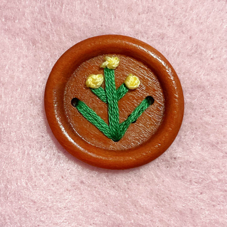 A brown 9-hole button with green and yellow thread resembling a plant design on a pink textured background.