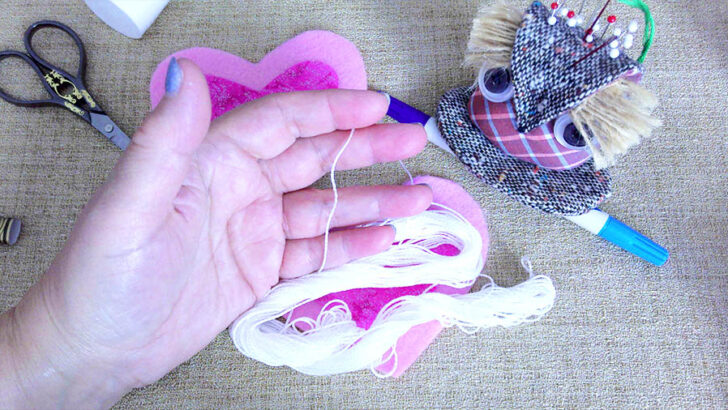 A hand holding white embroidery thread is in the foreground. Nearby are heart-shaped fabrics, scissors, and an owl pincushion on a burlap surface.