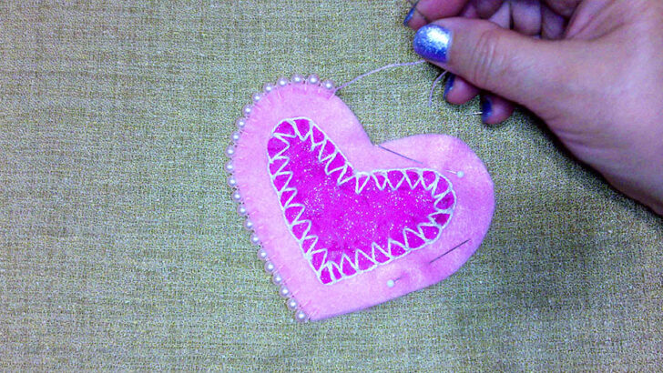 A hand holds a pink felt heart ornament decorated with white zigzag stitching and pearl beads along the edge