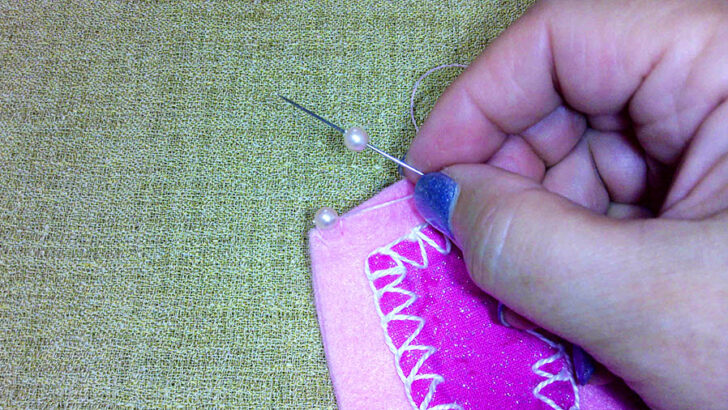 A hand sews pink fabric using a needle and thread on a textured green surface