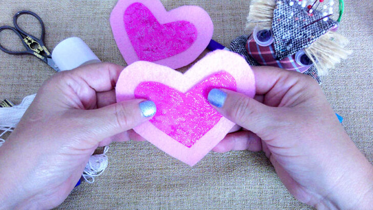 A pair of hands is shown holding a layered heart ornament. The larger heart is made of light pink felt, and a smaller magenta cotton heart with silver sparkles is centered on top. The pieces are securely glued together. In the background, tools like embroidery thread, scissors, and a playful pincushion are visible, hinting at the ongoing crafting process.