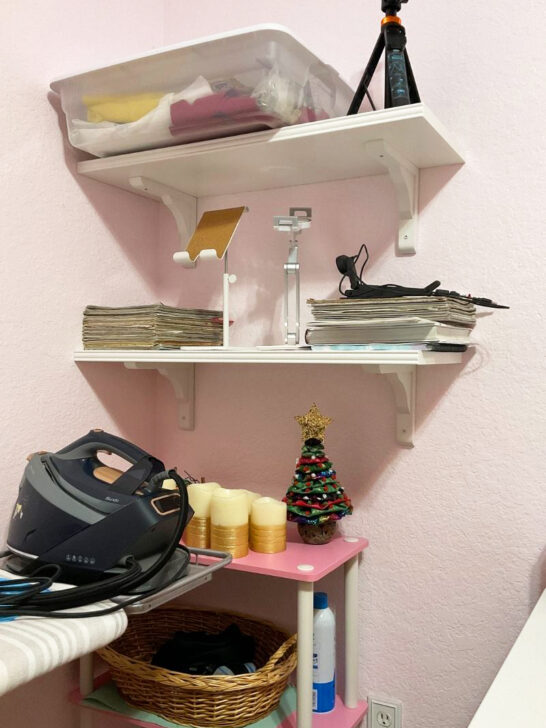 A pink sewing room featuring a sewing machine, ironing board, and a decorative shelf filled with sewing supplies