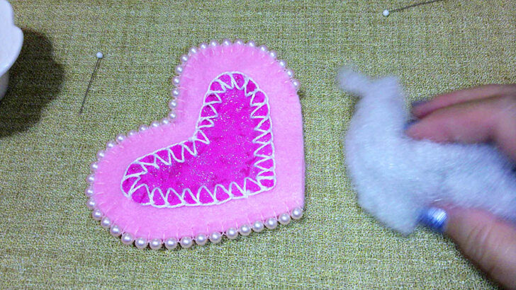 Close-up of a pink felt heart craft with a zigzag pattern, bordered by pearls. A hand is holding fluffy stuffing beside it