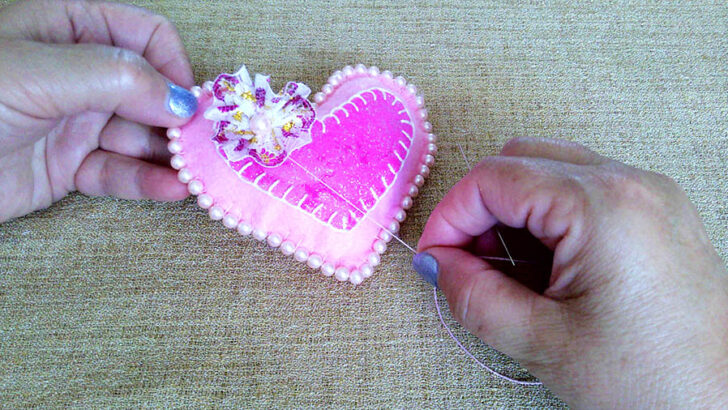 Hands are sewing a pink heart-shaped cushion adorned with pearls and a decorative flower.