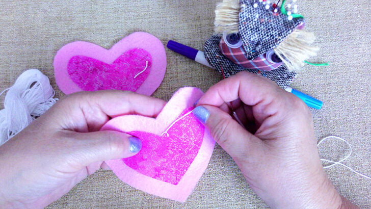 Hands are sewing two felt hearts, one pink and one purple, on burlap.