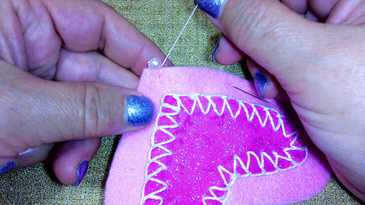 Hands sew a pink felt heart with white stitching on a textured surface