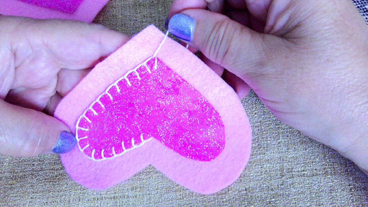 Hands sewing a bright pink glittery heart onto a light pink felt shape using white thread.