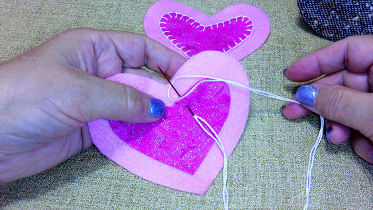 Hands sewing two pink felt hearts with a needle and white thread on a textured surface.