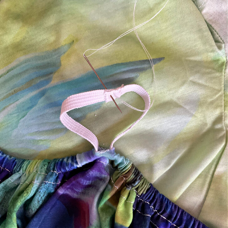 Close-up of sewing the ends of elastic on a colorful knit fabric. A needle with white thread is shown stitching the overlapping ends of a white elastic band, which has been threaded through the fabric casing to create a gathered effect. The fabric features a vibrant floral and watercolor design.