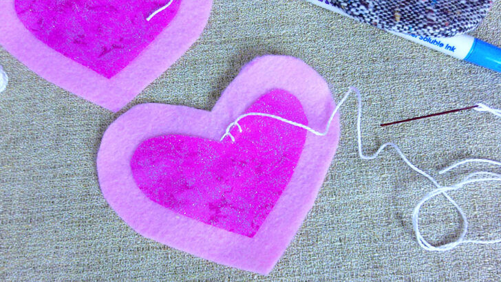 Two pink felt hearts on burlap are partially stitched with white thread.