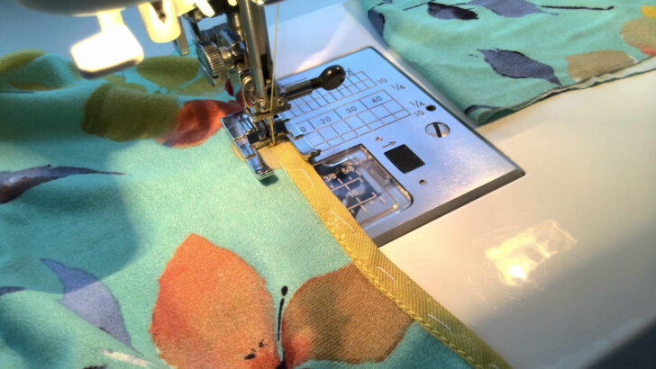 A sewing machine is shown stitching a piece of light green fabric with a colorful floral pattern. The machine uses an open-toe presser foot for topstitching, guiding a yellow bias tape along the fabric edge. The sewing needle is piercing through the bias tape as the stitch is being sewn. Bright lighting highlights the sewing process.