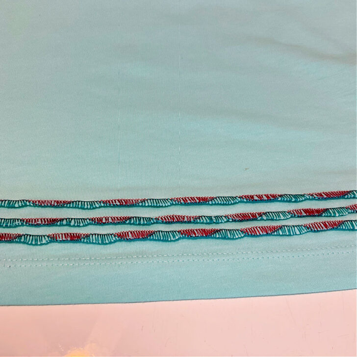 Close-up of a light green t-shirt hem decorated with three rows of wave stitches created using a serger. The stitches feature contrasting green and burgundy threads, providing a decorative and textured finish. The original hem remains visible below the decorative rows, highlighting the stitching technique.
