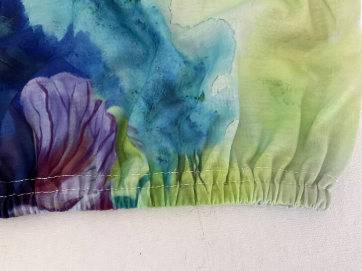 Close-up of a t-shirt hem with elastic inserted to create a gathered effect. The vibrant knit fabric features a watercolor-style design with blue, green, and purple tones. The gathers along the hem add a sporty and decorative touch to the garment.