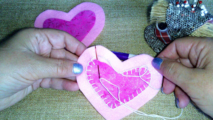 hands sew a small pink felt heart onto a larger pink felt heart using a needle and thread.