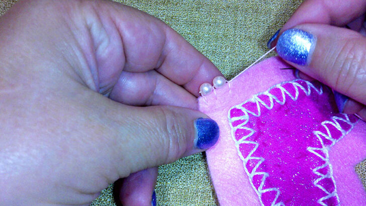 hands sew beads onto pink felt with zigzag stitching on a sparkly pink heart