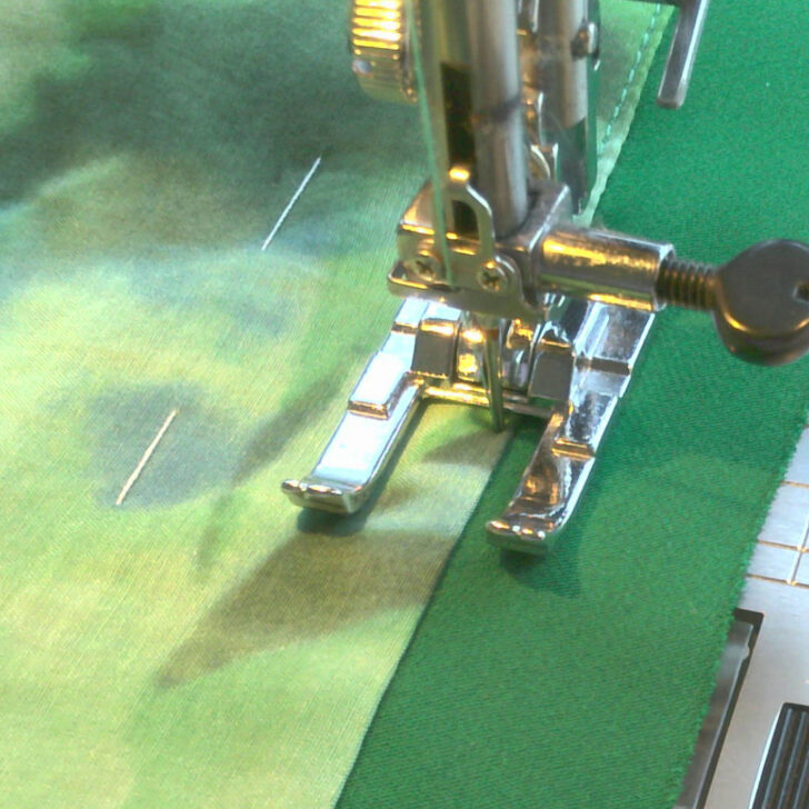 A sewing machine is shown with an open-toe presser foot stitching along the edge of two green fabric pieces. The needle is positioned at the seam, and the open-toe foot allows for clear visibility of the stitching path. The fabrics are aligned with visible pins holding them together. The presser foot and needle mechanism are in focus, while the fabric’s texture and seam are highlighted.