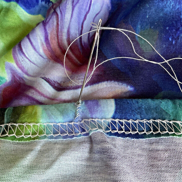 Close-up of hand sewing to close a hole in the hem of a colorful knit fabric. A needle threaded with white thread is shown stitching the small opening, which was used to insert elastic into the hem casing. The vibrant fabric features floral patterns, and the serged edge of the hem is visible.