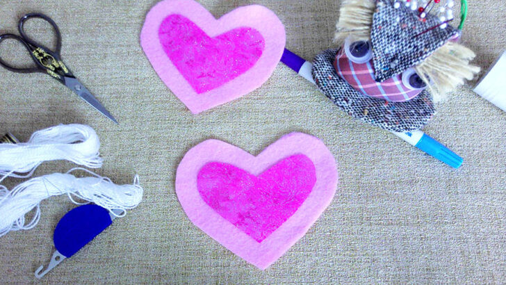 The image showcases two large felt hearts in pastel pink, with smaller, glittery magenta cotton hearts positioned on top. Scissors, embroidery thread, and a needle threader are nearby, hinting at the next steps in sewing a felt heart ornament