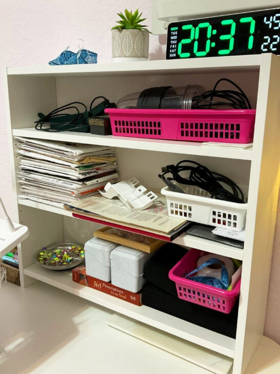 white desk displaying a laptop, a clock, and a basket containing assorted sewing supplies and decors