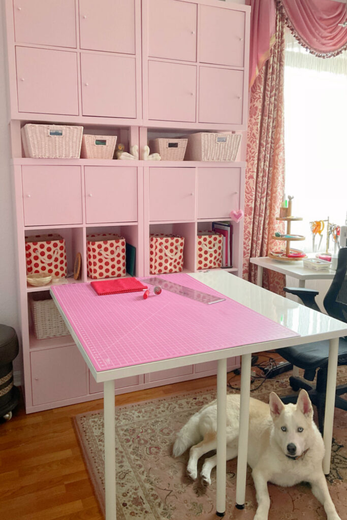 Pink-themed IKEA Kallax craft room setup with organized shelves, decorative bins, and a white crafting desk with a pink cutting mat. A white dog rests comfortably underneath the desk.
