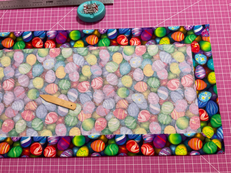 The image shows the full backing fabric with two mitered corners. A seam press tool and a magnetic pin holder with pins are also visible.