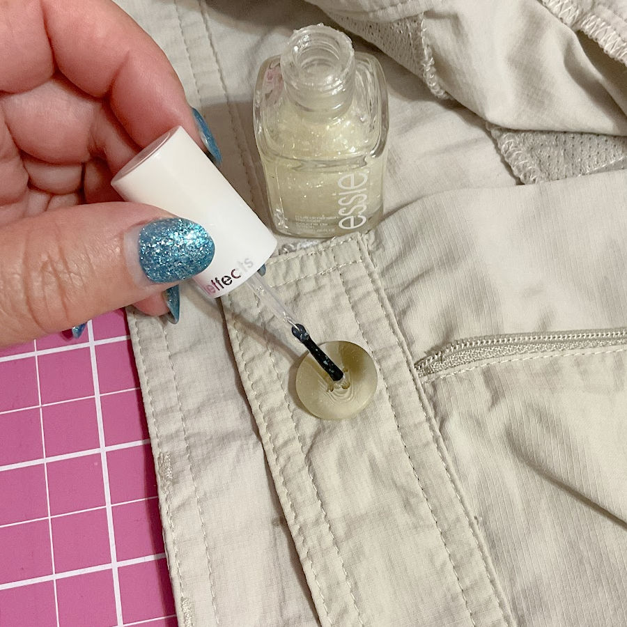 A woman's hand with a nail polish bottle placing a tiny drop of nail polish on the thread from a button to avoid unravelling