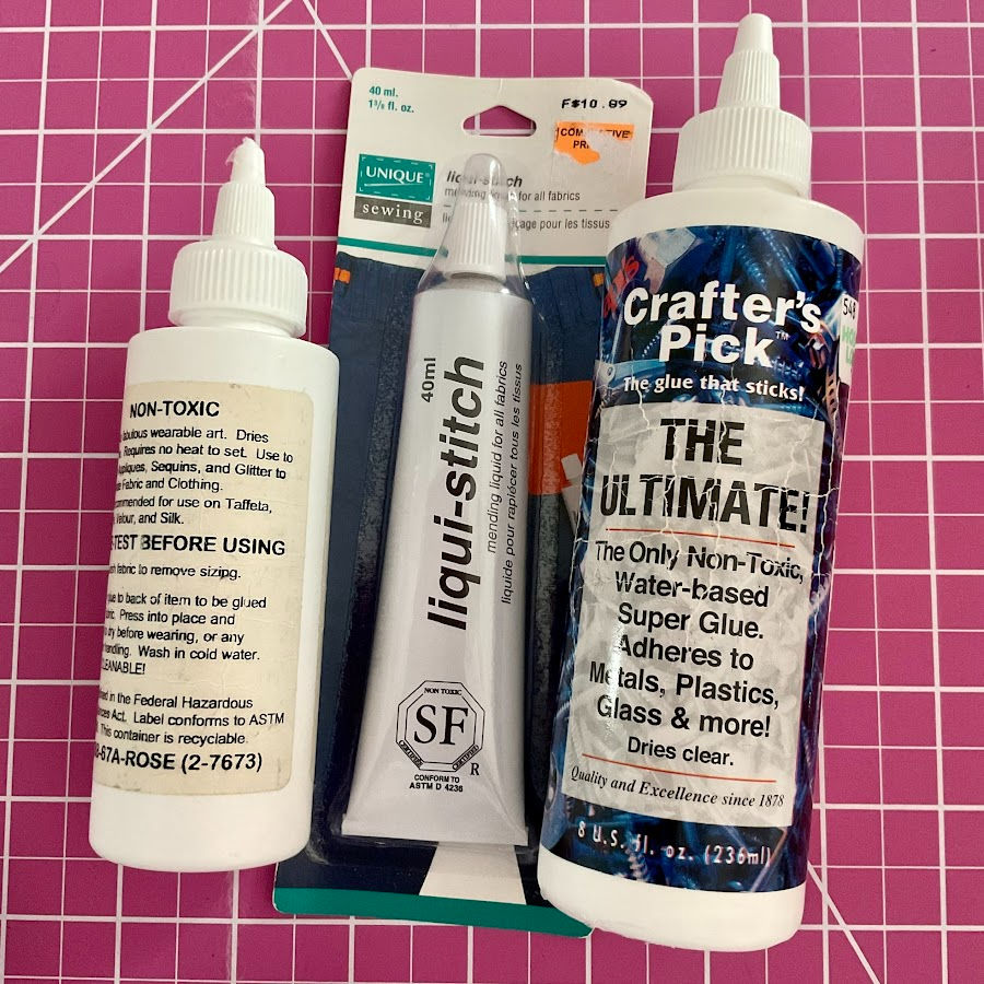 Three different tubes of fabric glue on display on a pink cutting mat