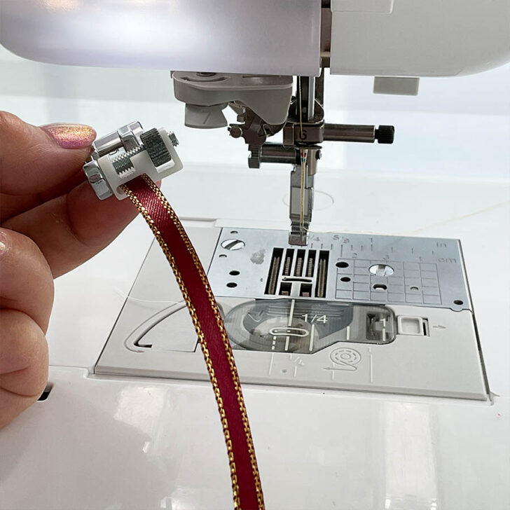 A woman's hand holding a sequin presser foot before attaching it to the sewing machine. A sewing machine in the background.