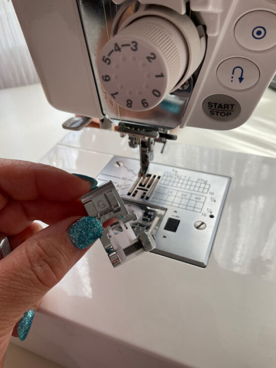 A woman's hand holding a sequin presser foot before attaching it to the sewing machine. A sewing machine in the background.