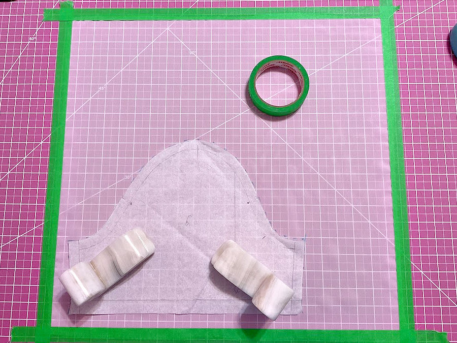 A piece of chiffon material affixed to the cutting mat with painter's tape, a roll of painter's tape and a pattern piece held by two stone weights