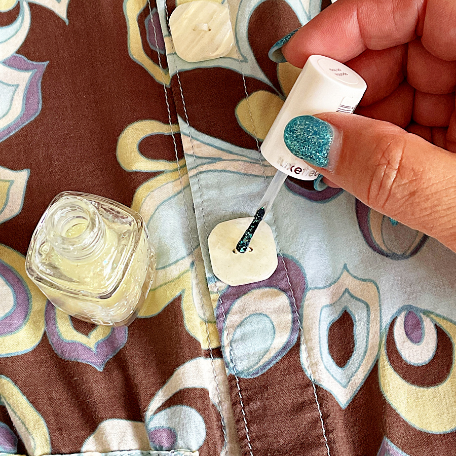 Close-up of a person applying clear nail polish to the center of a button on a patterned shirt. A small bottle of polish sits nearby on the fabric.