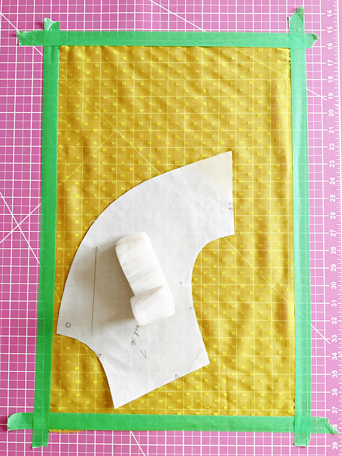 Yellow sheer fabric secured to a pink cutting mat using green painter's tape, with a paper pattern piece and fabric weight placed on top.