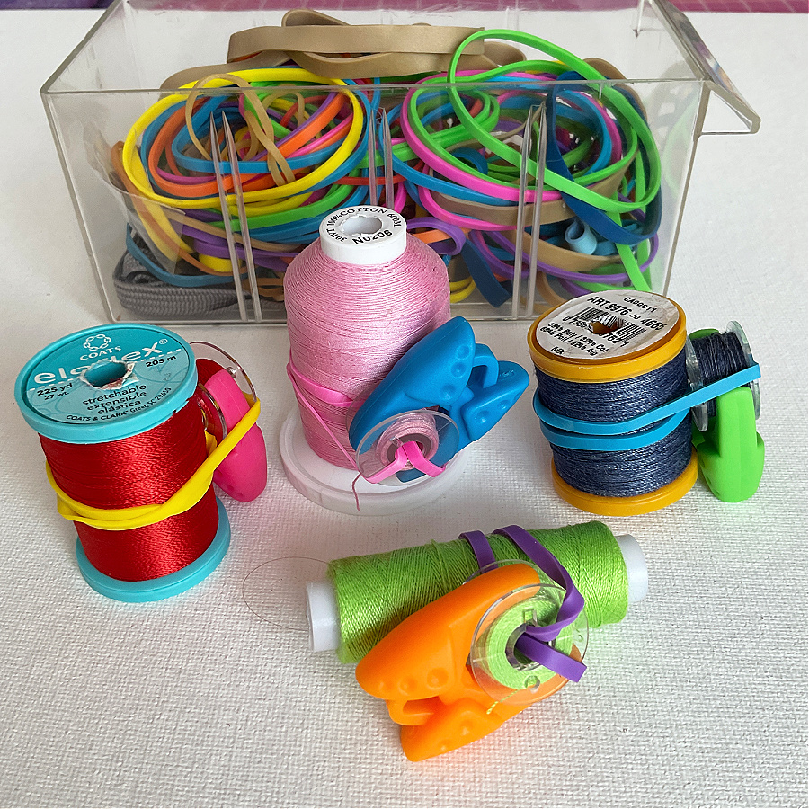 Colorful rubber bands stored in a clear plastic container. Several spools of thread are shown with rubber bands and plastic clips wrapped around them to keep loose thread ends in place.