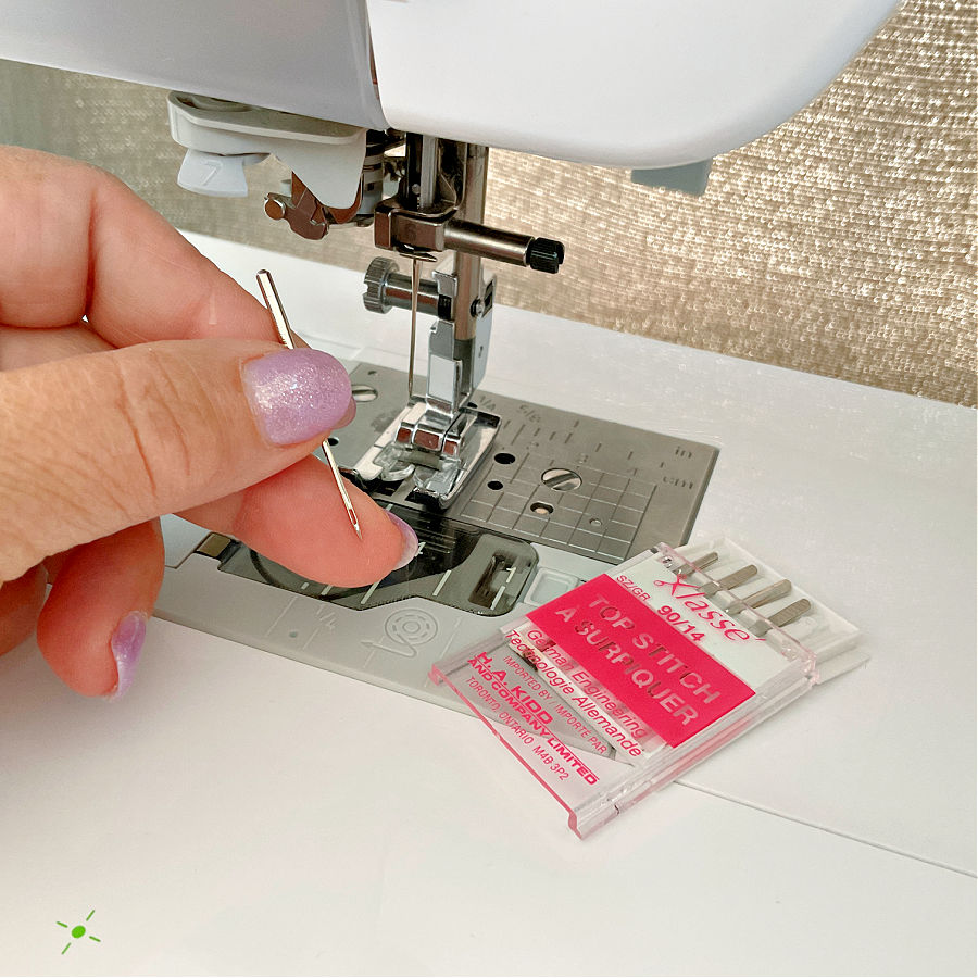 a woman's hand holding a needle near a sewing machine needle assembly while a klasse needle pack is visible on the machine