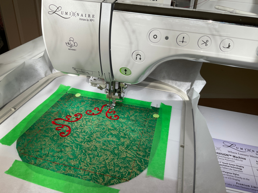 Embroidery machine stitching a red decorative design on green fabric. The fabric is secured to the stabilizer using green painter's tape instead of hooping.
