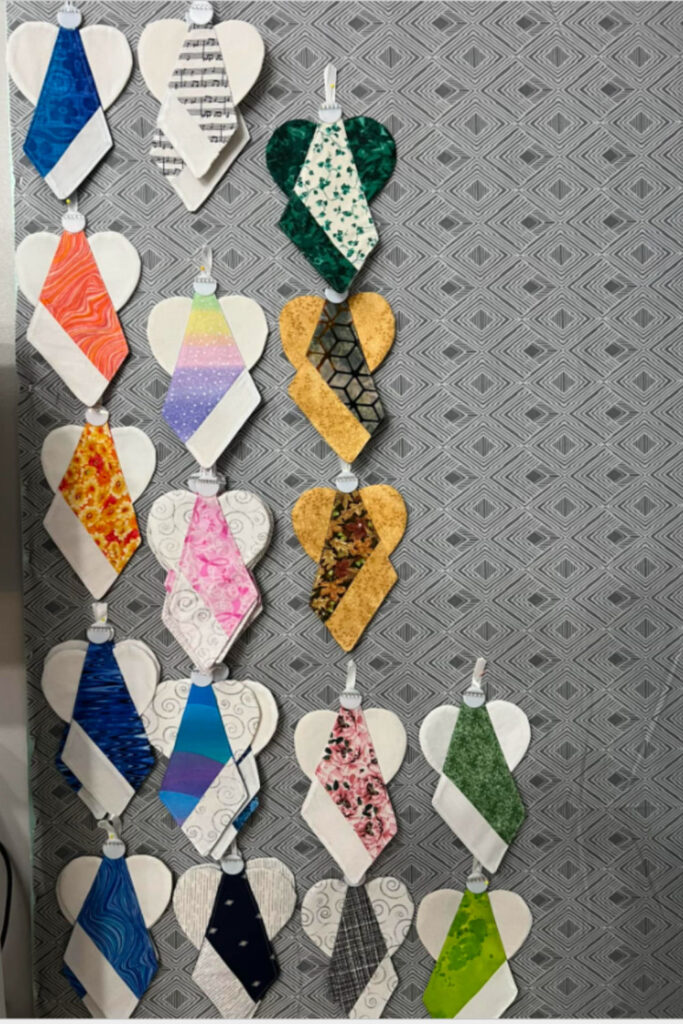 Handmade fabric angel ornaments arranged in rows on a gray geometric background. Each angel features a variety of colorful printed fabrics for the body and white fabric wings, with small loops for hanging. The angels are displayed flat and evenly spaced, showcasing a mix of patterns and textures.