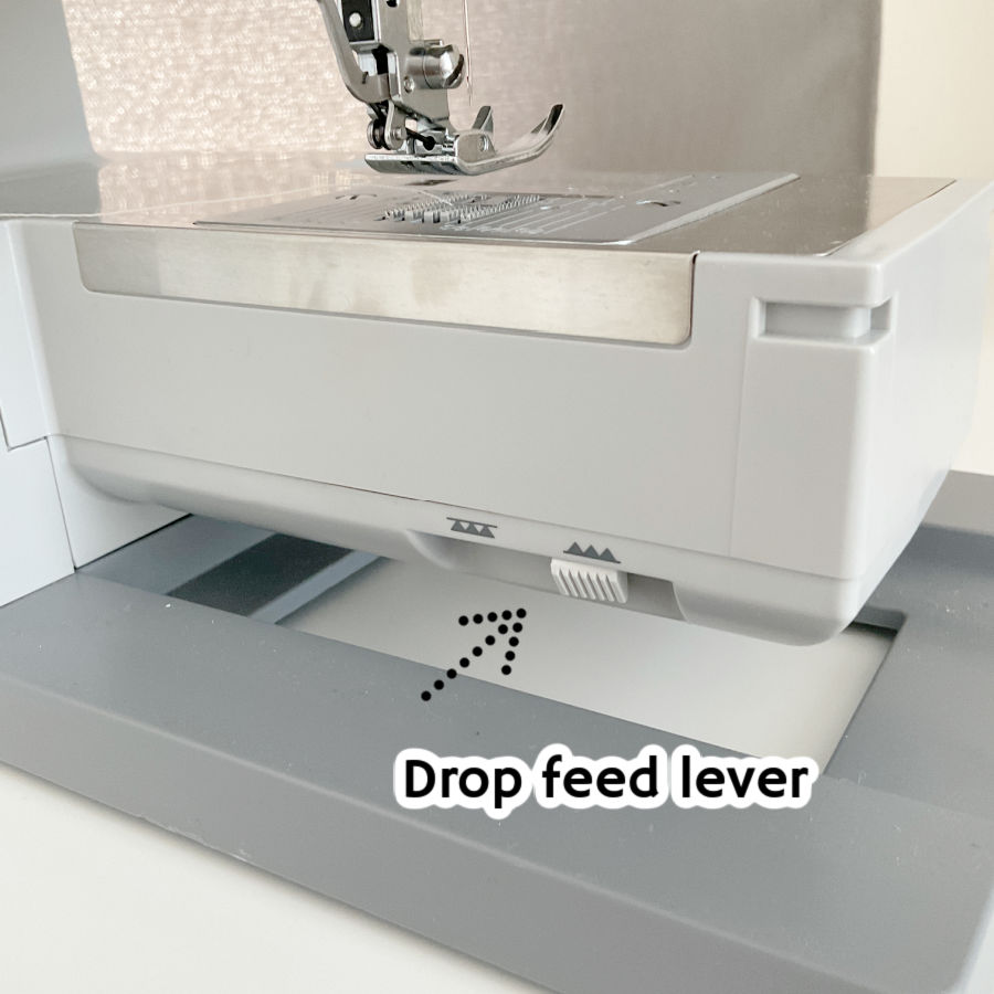 the bottom of a sewing machine with the drop feet lever highlighted by an arrow pointing at it