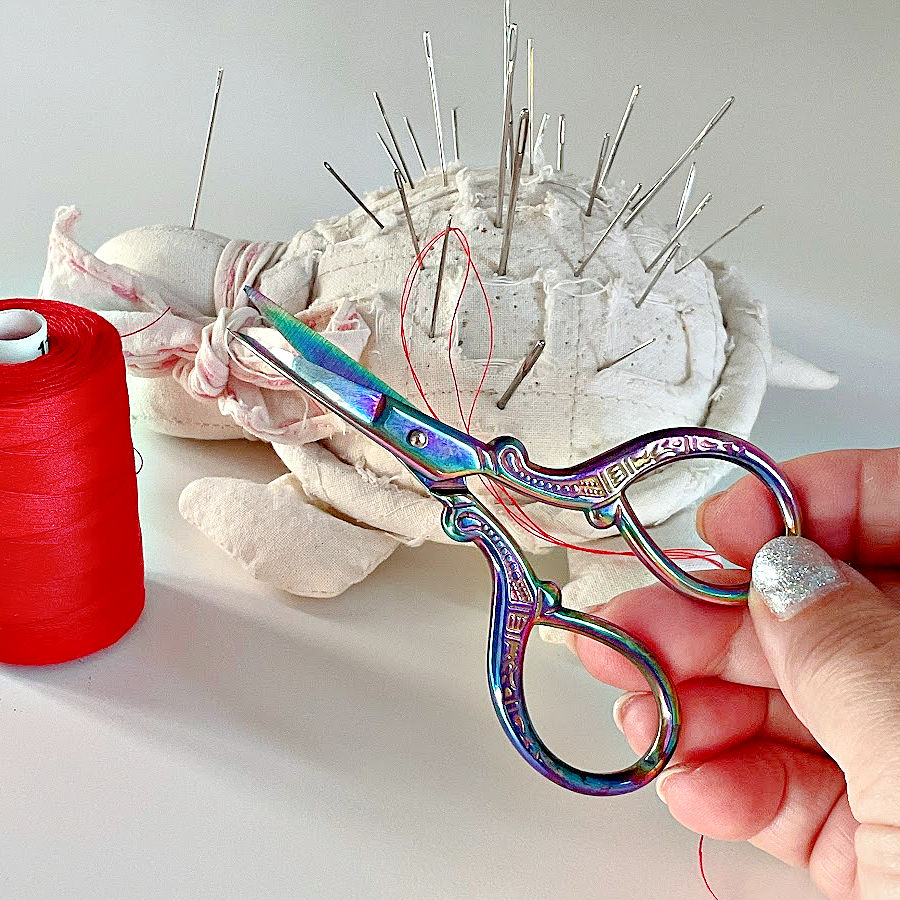 a womans hand holding a pair of small embroidery scissors a pincushion with needles and a spool of thread in the background