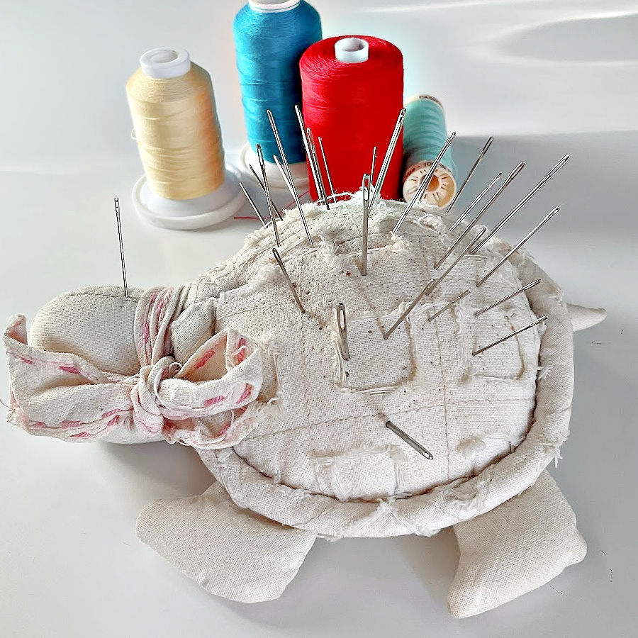 multiple hand sewing needles og various sizes in a pincushion shaped like a turtle four thread spools in the background