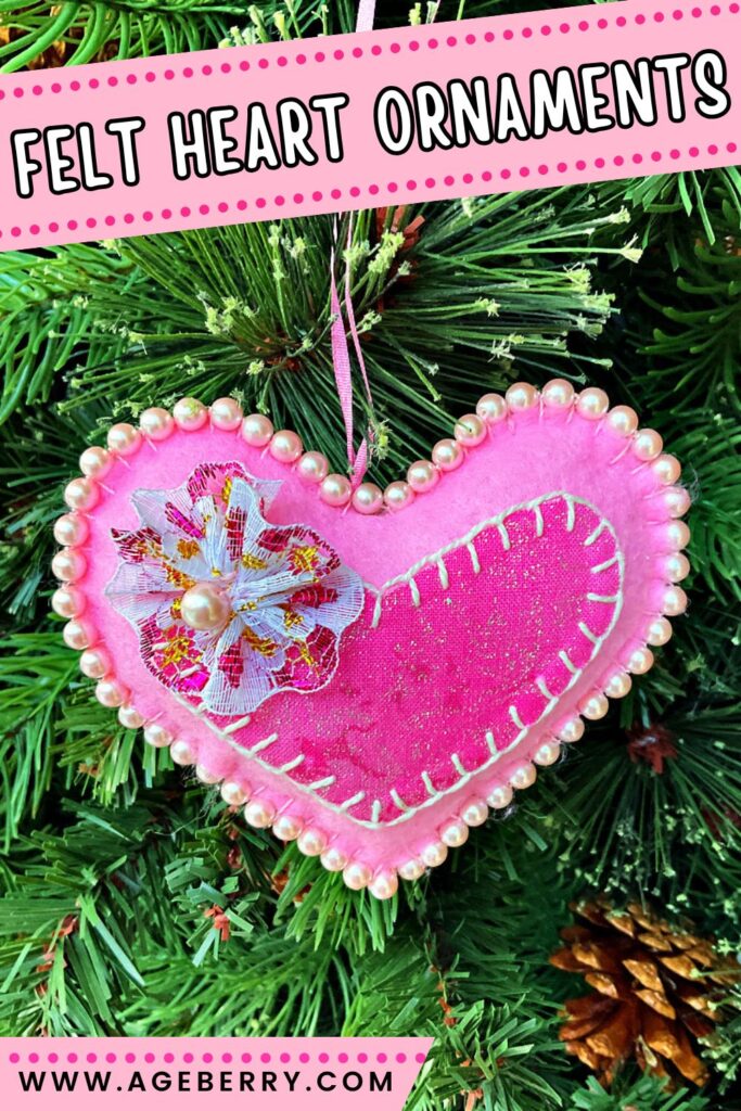 Handmade pink felt heart ornament decorated with blanket stitching, pearl beads around the edges, and a fabric flower with a pearl center. The ornament is hanging on a green Christmas tree.