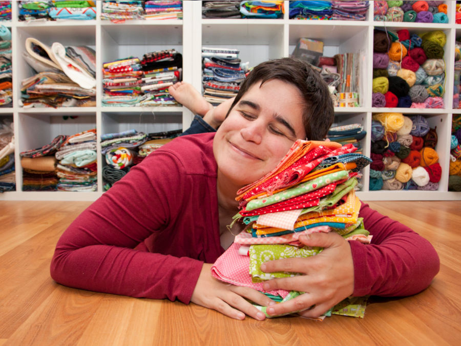 a woman laying on her belly on the floor hugging a pile of multi colored fabric a fabric storage unit in the background