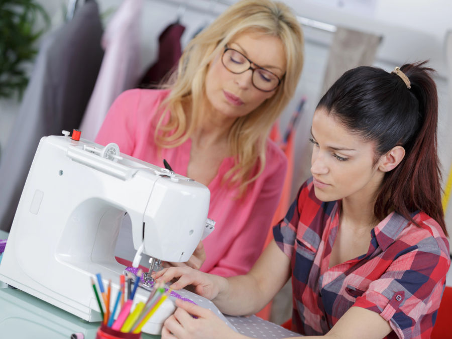 two women in front of a sewing machine cooperating at work one woman reads a notebook while the other woman is sewing a stitch