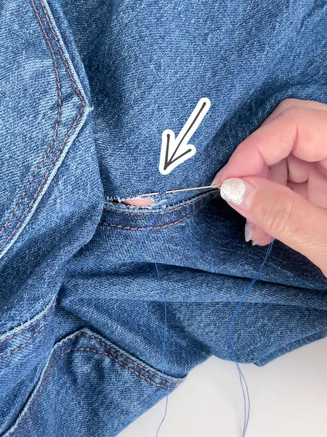 image shows a womans hand inserting a needle to start the blanket stitch to mend the rip in the jeans