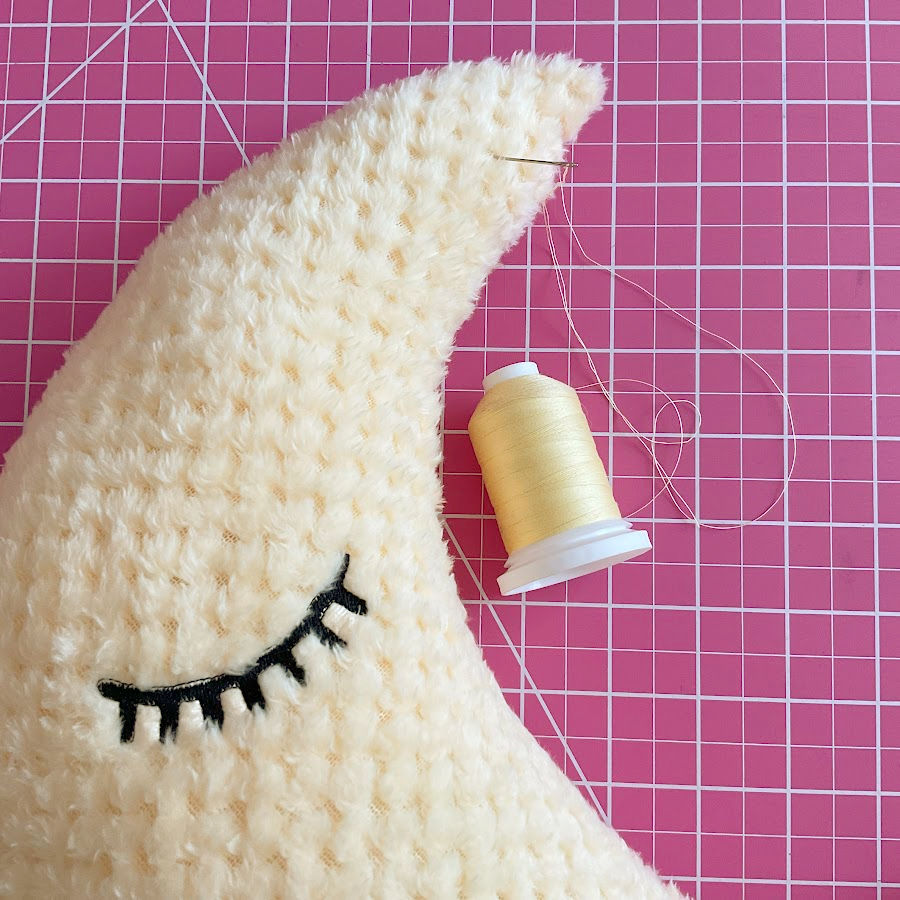 Detail image of the top of the moon toy while the opening left for stuffing is being closed by hand a threaded needle and a spool of thread are also visible