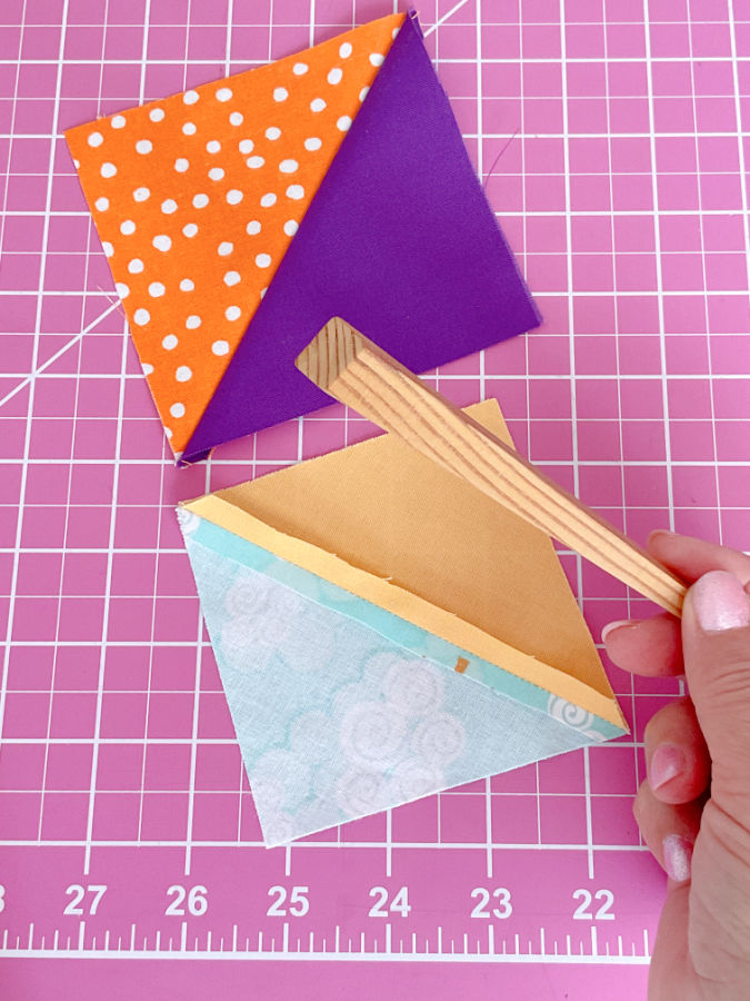 two half square triangles one right side up the other wrong side up and a womans hand holding a wooden finger presser tool