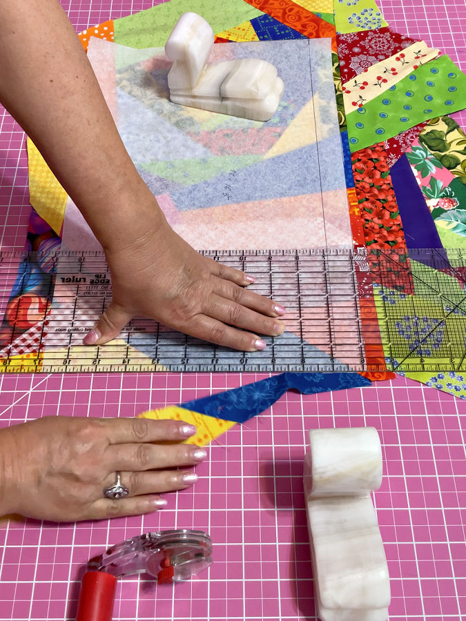 a womans hands shown while cutting a rectangular panel from a crumb block using a quilting template two stone weights and a rotary cutter are visible