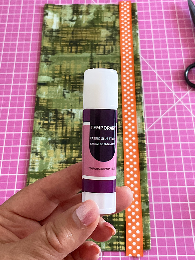 a womans hand holding a tube of temporary fabric glue a piece of material on a cutting table in the background