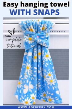Blue floral kitchen towel hanging from a stove handle using a plastic snap closure. The towel is wrapped around the stove rod and fastened at the front with a pink snap, creating a secure hanging towel without sewing. The image shows how snaps can replace traditional hanging towel holders