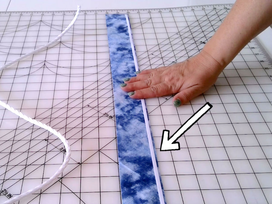 using wonder tape to hold together the fabric folded over the cord before sewing a womans hand shown pressing a strip of wonder tape to the edge of the fabric strip the cord to be wrapped near the strip